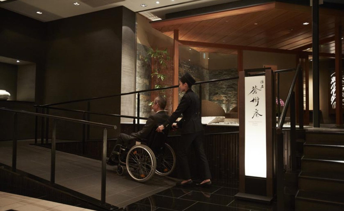 Staff assists guest in wheelchair up a sleek indoor ramp at Keio Plaza Hotel. Accessible rooms feature a roll-in shower.