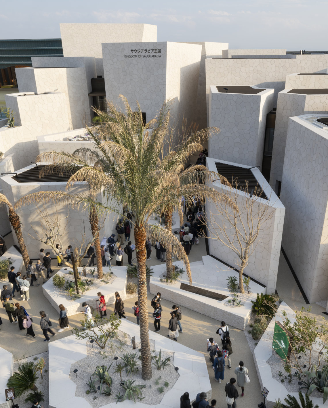 Visitors walk through a courtyard with palm trees and modern white stone buildings at the Saudi Arabia Pavilion, Expo 2025.