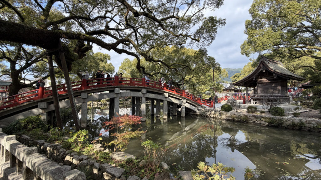 Arched red bridge over a pond at Dazaifu Tenmangu, surrounded by trees and stone structures in a scenic, tranquil shrine setting.