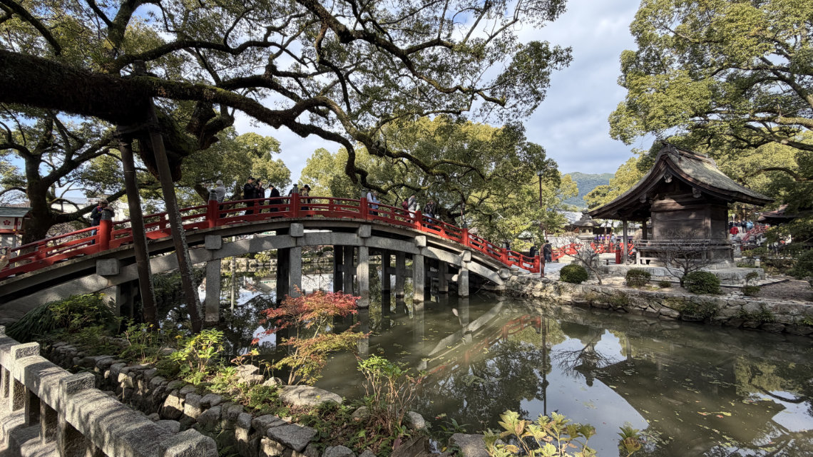 Arched red bridge over a pond at Dazaifu Tenmangu, surrounded by trees and stone structures in a scenic, tranquil shrine setting.