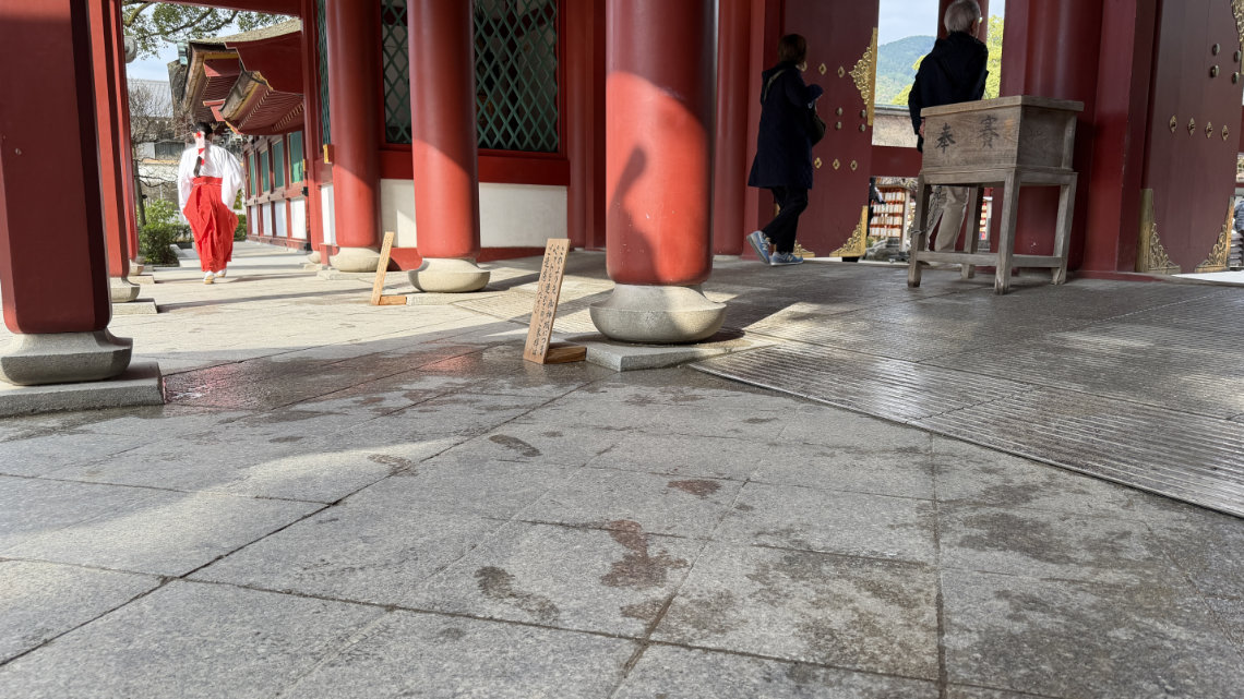 Red pillars at Dazaifu Tenmangu with a slope for accessibility, wet floor signs, and visitors near a wooden offertory box.