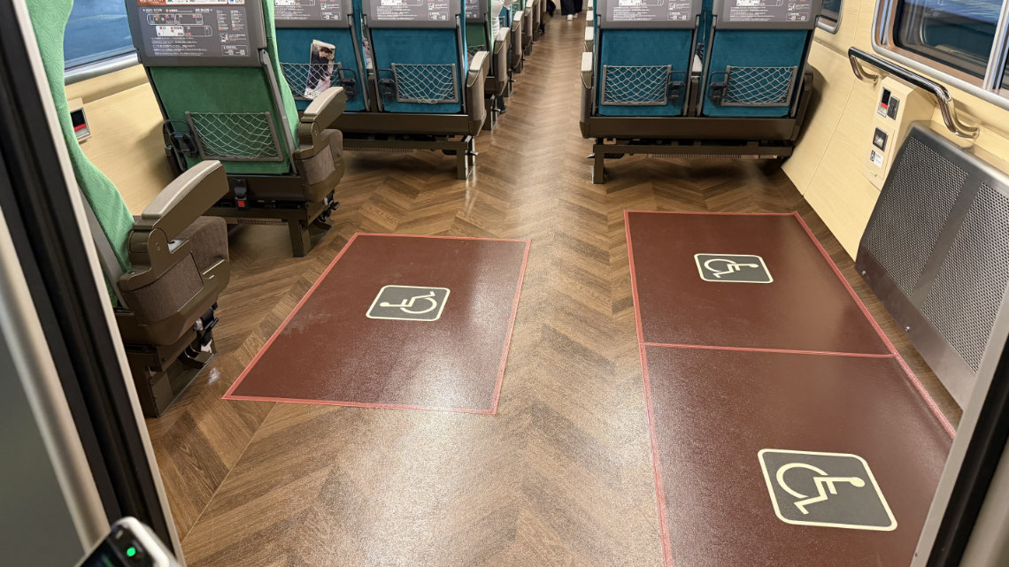 A train car interior showing two dedicated wheelchair spaces marked with large white symbols on the wood-grain floor, located adjacent to green passenger seats.