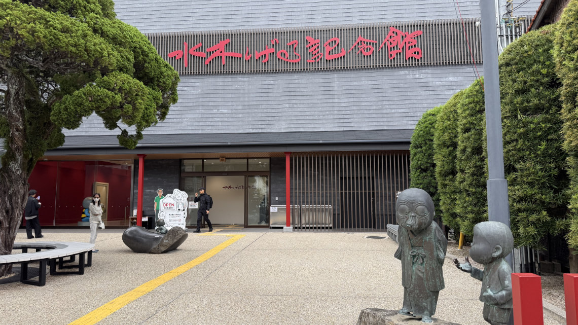 The Mizuki Shigeru Museum entrance featuring red Japanese signage, automatic sliding doors, yellow tactile paving, and bronze statues of yokai characters in the foreground.