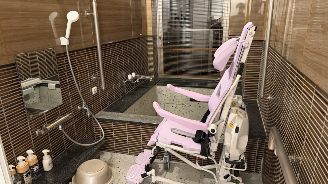 A tiled bathroom featuring a deep Japanese soaking tub and a specialized pink mechanical lift chair. A shower station with toiletries and a mirror is visible on the left.