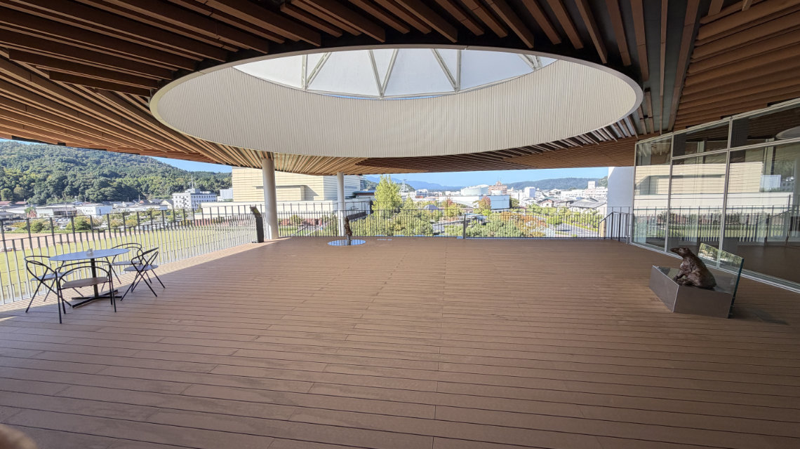 A spacious wooden observation deck with a large circular roof opening, offering a view of distant mountains. It features a table, chairs, and small animal sculptures.