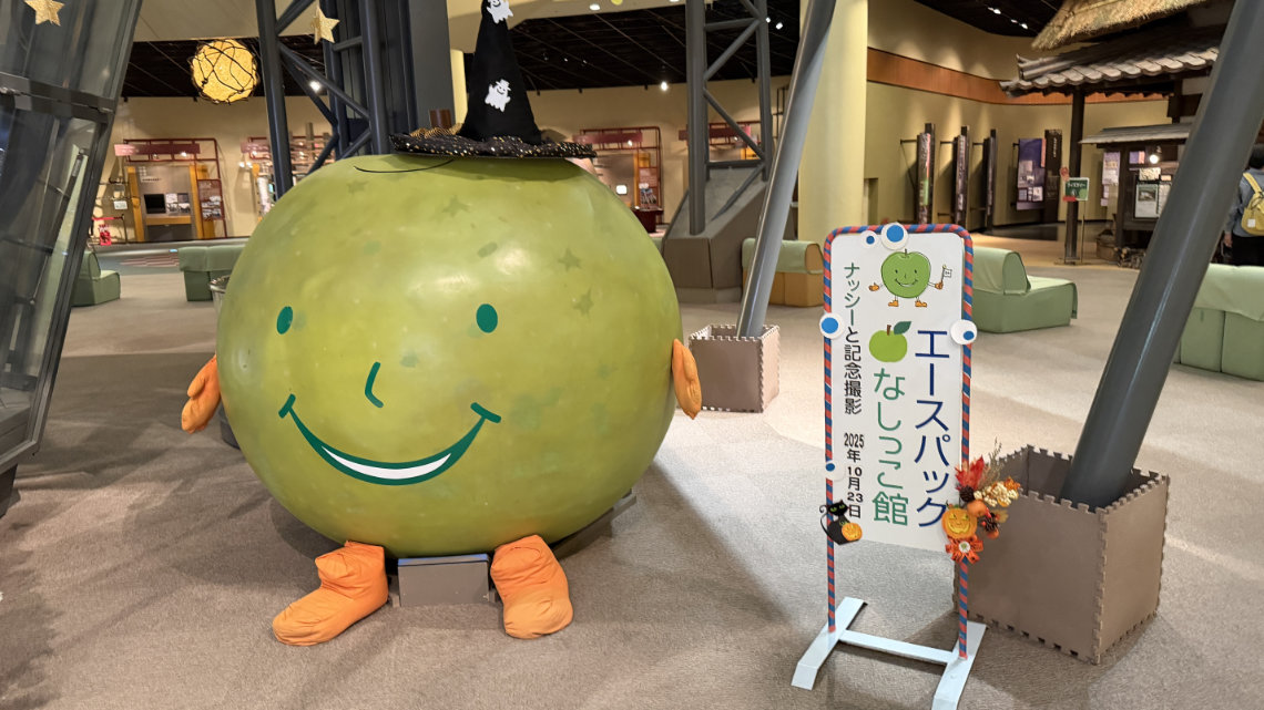 A large, round, green pear mascot named Nassy wearing a black witch's hat, standing next to a photo signboard inside a museum.
