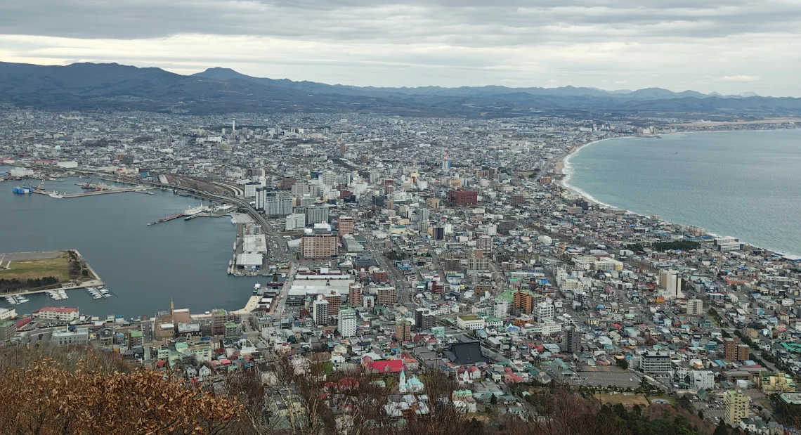A panoramic view of the city of Hakodate from the summit of Mt. Hakodate on a cloudy day, showing the city's unique shape, the busy harbor on the left, and the open ocean on the right.