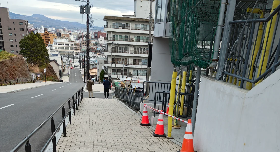 A view looking down a steep, paved street in a hilly area of Hakodate. There is a handrail on the left side and a construction site with scaffolding on the right. Two people are standing at the top of the slope, looking down towards the city and distant mountains under a cloudy sky.