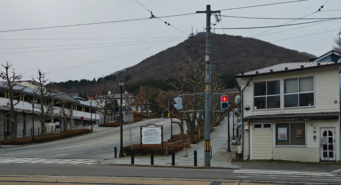 A wide cobblestone street sloping upwards towards Mt. Hakodate, which has a tower on its peak. A red traffic light is visible on a utility pole, and there is a white wooden building on the right side of the street.