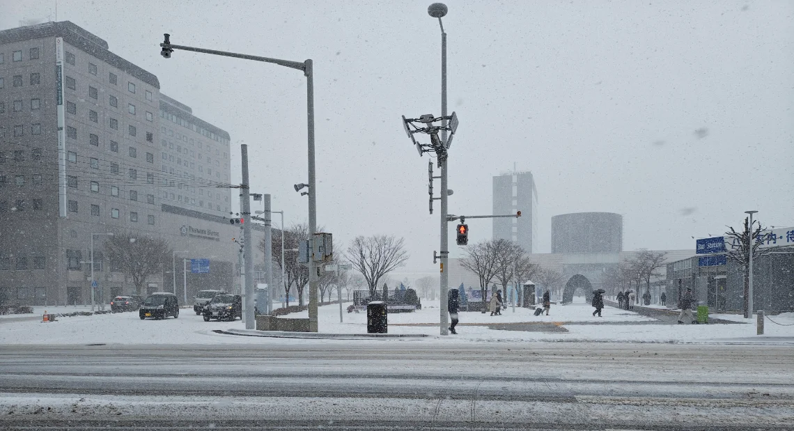 A snowy street scene at a large intersection in Hakodate during a heavy snowfall. The ground is covered in snow and slush, and pedestrians are walking on the sidewalks. A bus stop shelter is visible on the right, and a large hotel building is on the left.