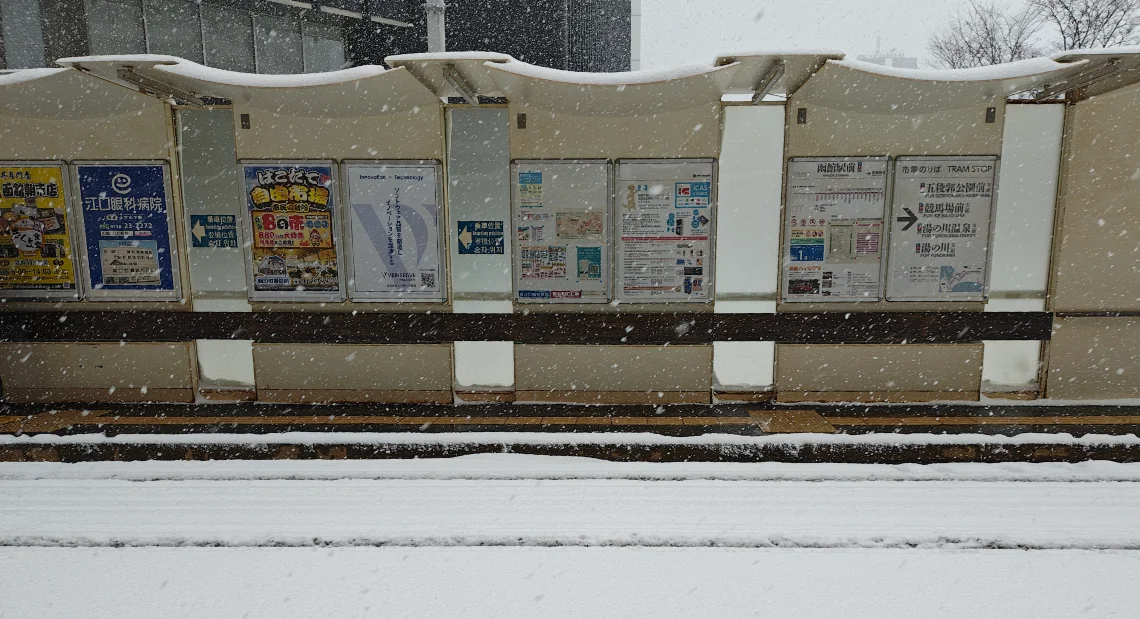 A tram stop shelter in Hakodate covered in fresh snow while snow is falling. Timetables and advertisements are visible on the back panel of the shelter, and the tram tracks in the foreground are blanketed in white.